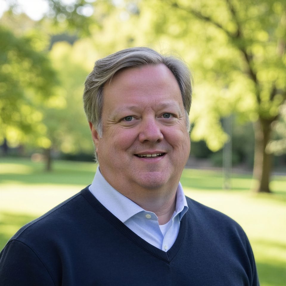 John David Marx, founder of Webspinner, smiling outdoors in a navy sweater over a light blue shirt, trees in soft focus behind him.